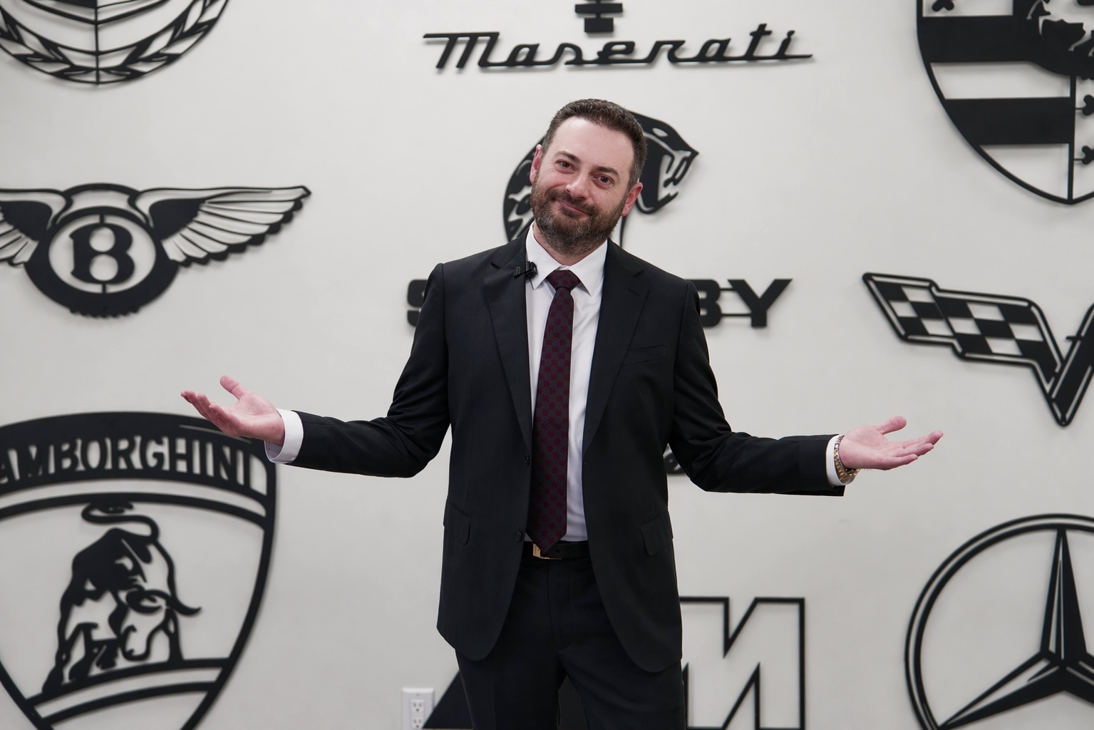 Harry H. Terzian standing in front of a wall of major luxury auto manufacturer logos, Maserati, Bentley, Lamborghini, Mercedes, Porsche, Shelby, Corvette